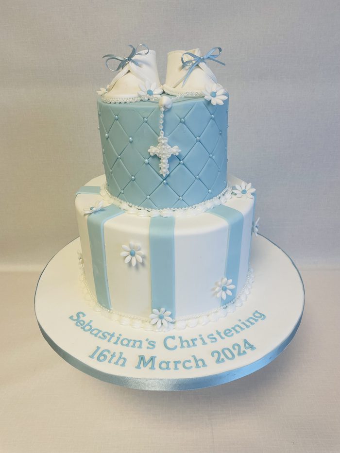 Two tier blue and white christening cake with sugar bootees, a cross and daisy detail