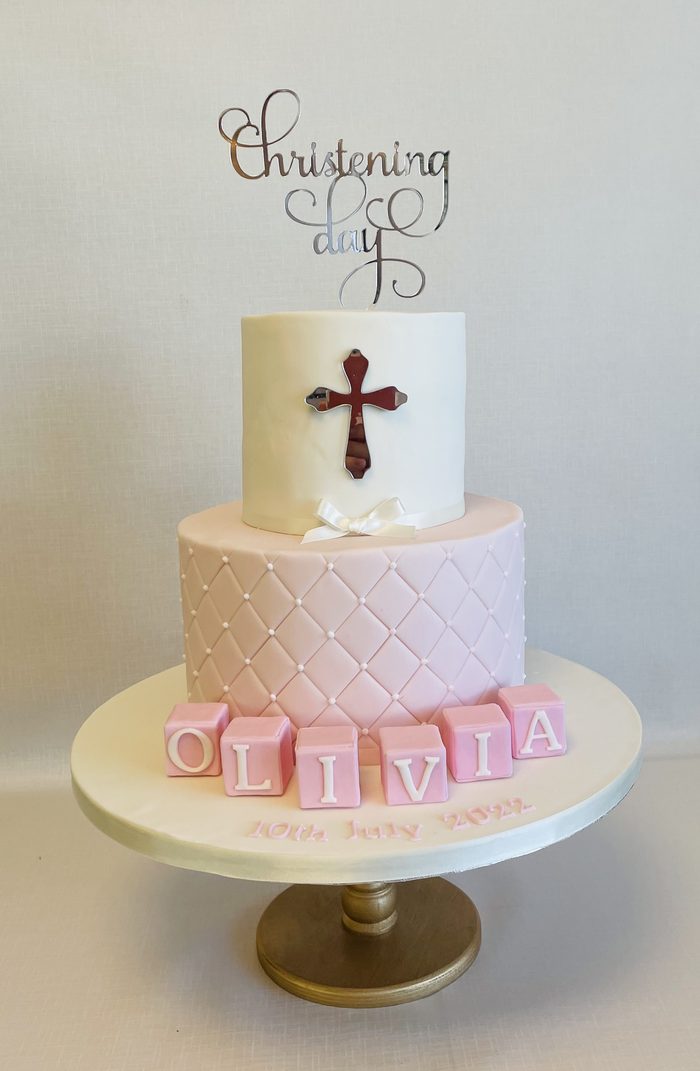 Two tier pink christening cake with a quilted lower tier, cross topper and name blocks spelling Olivia